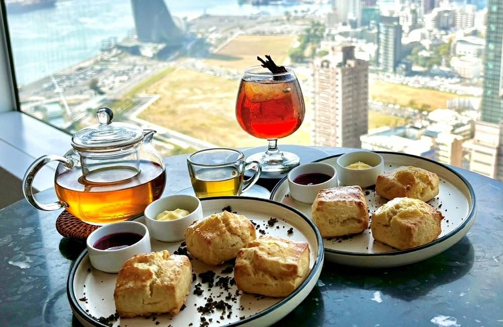 SEVENTEEN小分隊cxm演唱會高雄開唱懶人包整理 - Travel x Freedom 旅誌字遊 threeonelee.com 高樓層窗邊桌面擺放雙人下午茶點心與茶飲,背景為高雄市區街景與港灣視角的實際用餐畫面